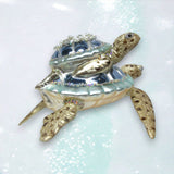 The December Diamonds ORN: Blue Turtle w/Baby on Back features a sparkling blue and silver shell with beads and jewels, displayed on a shimmering surface.
