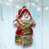ORN: Pirate Santa Captain by December Diamonds features a glass Christmas ornament of Pirate Santa holding a gold treasure chest adorned with jewels, displayed against a pale wood background with twinkling string lights above.