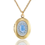 The Vintage Inspired Oval Light Blue Crystal Locket Necklace by Anne Koplik Designs, Inc. features a gold oval pendant with a light blue crystal center surrounded by pearls, creating an elegant keepsake for treasured photos.