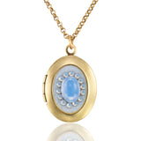 The Vintage Inspired Oval Light Blue Crystal Locket Necklace by Anne Koplik Designs, Inc. features a gold oval pendant with a light blue crystal center surrounded by pearls, creating an elegant keepsake for treasured photos.
