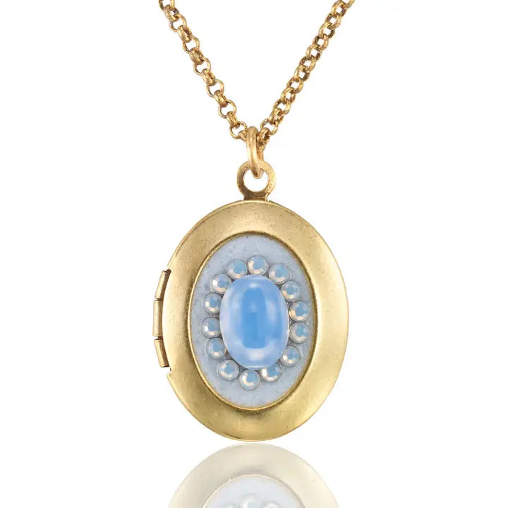 The Vintage Inspired Oval Light Blue Crystal Locket Necklace by Anne Koplik Designs, Inc. features a gold oval pendant with a light blue crystal center surrounded by pearls, creating an elegant keepsake for treasured photos.
