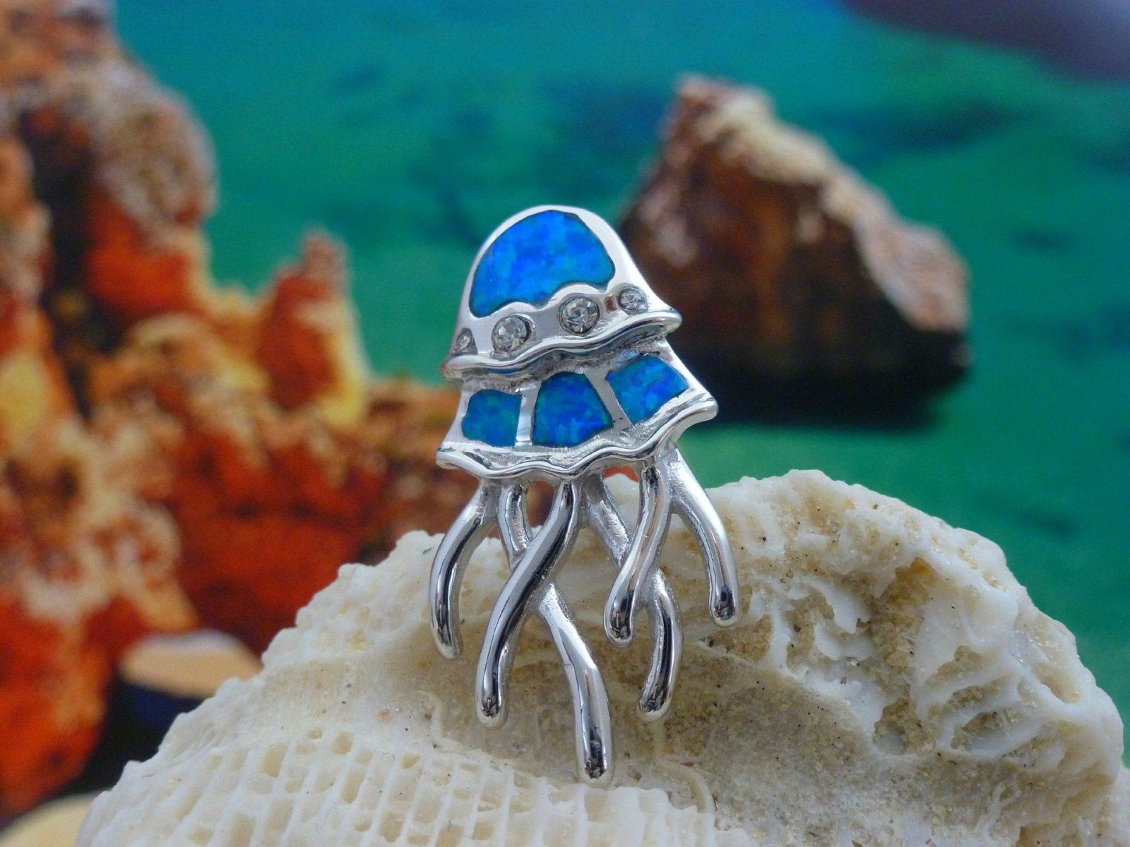 The Caribbean Opal Jellyfish-Pendant ONLY by Jewelry Network features a silver jellyfish design with blue synthetic opal inlays, displayed on a seashell with shimmering accents from softly blurred rocks and water in the background.