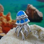 The Caribbean Opal Jellyfish-Pendant ONLY by Jewelry Network features a silver jellyfish design with blue synthetic opal inlays, displayed on a seashell with shimmering accents from softly blurred rocks and water in the background.