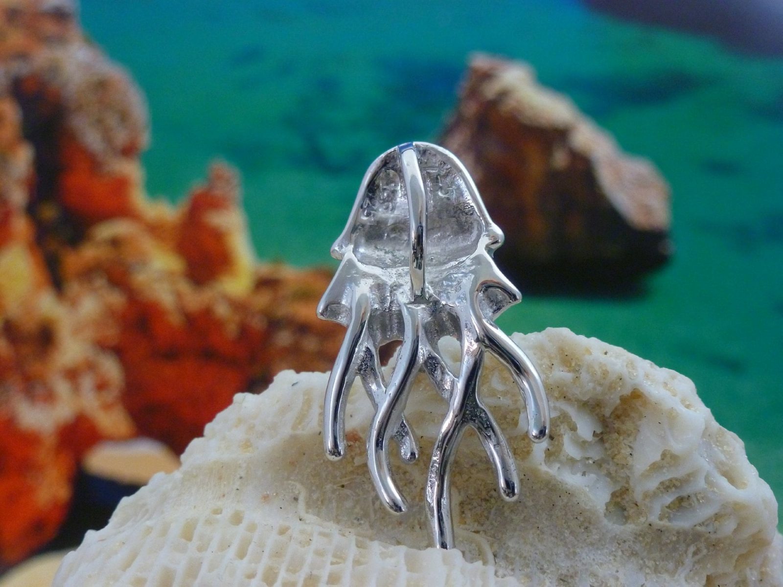 The Caribbean Opal Jellyfish-Pendant ONLY by Jewelry Network, crafted in 925 Sterling Silver with curved tentacles and a rhodium plate finish, rests on a textured white rock before a vibrant blue and orange seaside backdrop.