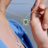 A person adjusts their Shanore Sterling Silver August Birthstone Turtle Necklace with green Swarovski crystals. They wear a blue garment against a softly blurred background.