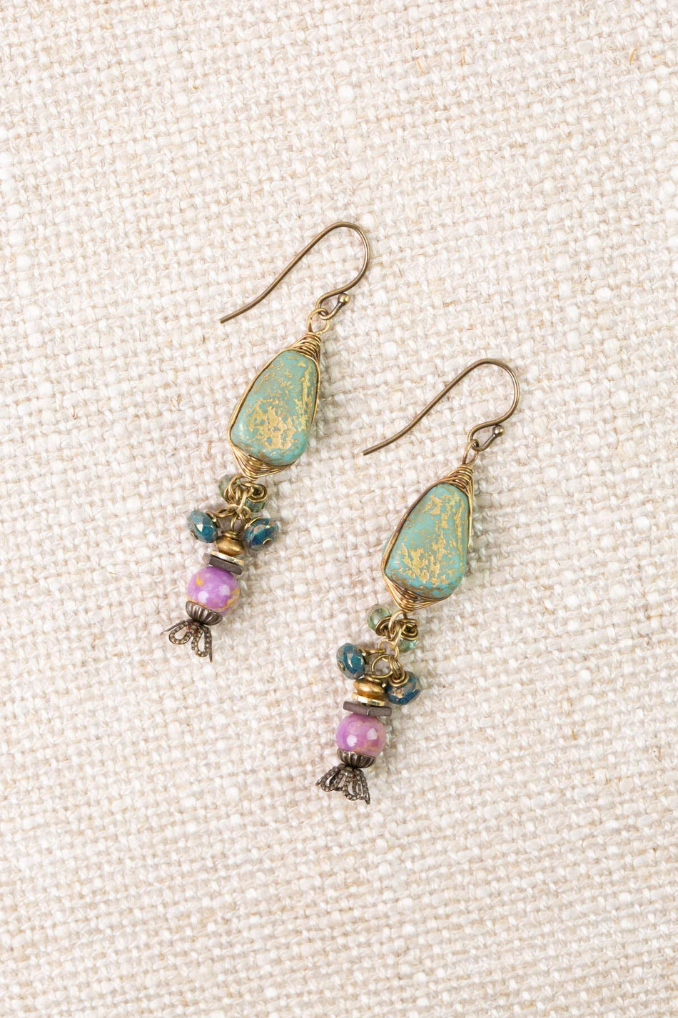 A pair of Anne Vaughan “ER: Folklore Phosphorite, Czech Glass Herringbone” handcrafted dangle earrings featuring green stone tops, blue and purple Czech Glass beads, antique brass accents, and fishhook ear wires, displayed on a textured beige fabric background.