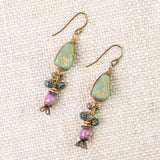A pair of Anne Vaughan “ER: Folklore Phosphorite, Czech Glass Herringbone” handcrafted dangle earrings featuring green stone tops, blue and purple Czech Glass beads, antique brass accents, and fishhook ear wires, displayed on a textured beige fabric background.