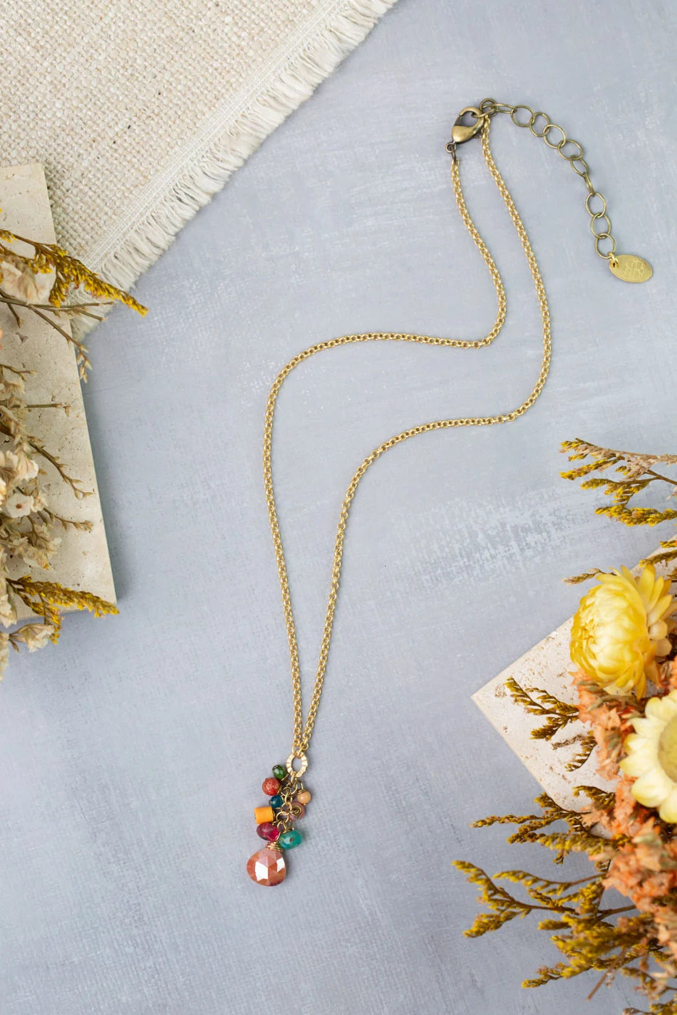 The Anne Vaughan NK: Farmers Market 15-17″ Coral, Sunstone, Czech Glass Cluster necklace is displayed on a light gray surface, surrounded by dried yellow and beige flowers and a piece of textured fabric. This gold necklace features colorful gemstone pendants, including vibrant Czech Glass accents.