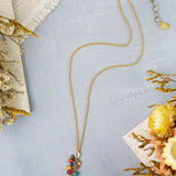 The Anne Vaughan NK: Farmers Market 15-17″ Coral, Sunstone, Czech Glass Cluster necklace is displayed on a light gray surface, surrounded by dried yellow and beige flowers and a piece of textured fabric. This gold necklace features colorful gemstone pendants, including vibrant Czech Glass accents.