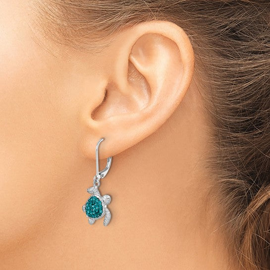 A close-up of a woman’s ear showcases Presco/Quality Gold’s Crystal Teal Turtle Dangle Earrings, with a green and silver shell design. Her pulled-back hair reveals the unique sterling silver accessory.