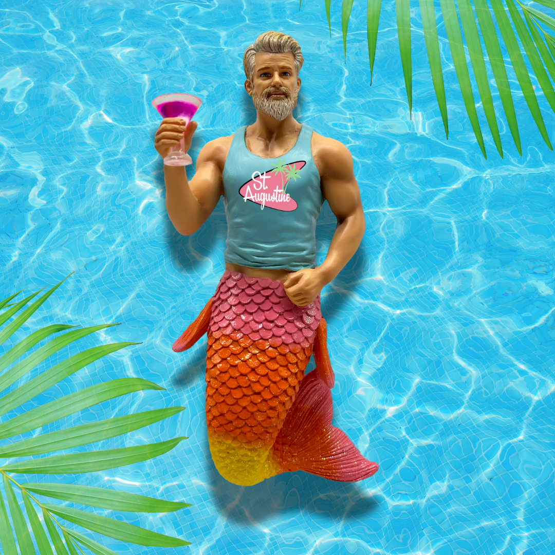 The Auggie Ornament by December Diamonds features a bearded merman in a light blue St. Augustine tank top, lounging with a pink drink amid hand-painted palm leaves—ideal for holiday decor or mermaid ornament collectors.