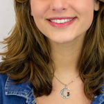 A smiling woman with wavy brown hair wears a denim jacket and the Crescent Moon w/Black Cat Necklace by Anne Koplik Designs, Inc., standing against a plain light background.