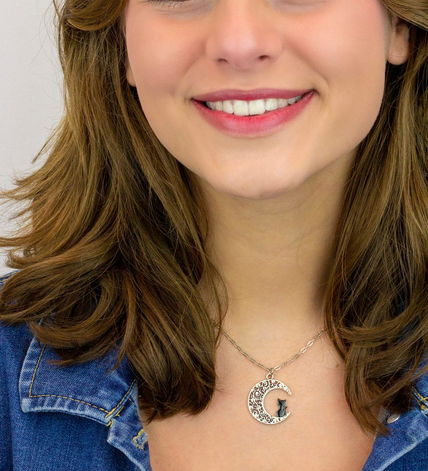 A smiling woman with wavy brown hair wears a denim jacket and the Crescent Moon w/Black Cat Necklace by Anne Koplik Designs, Inc., standing against a plain light background.