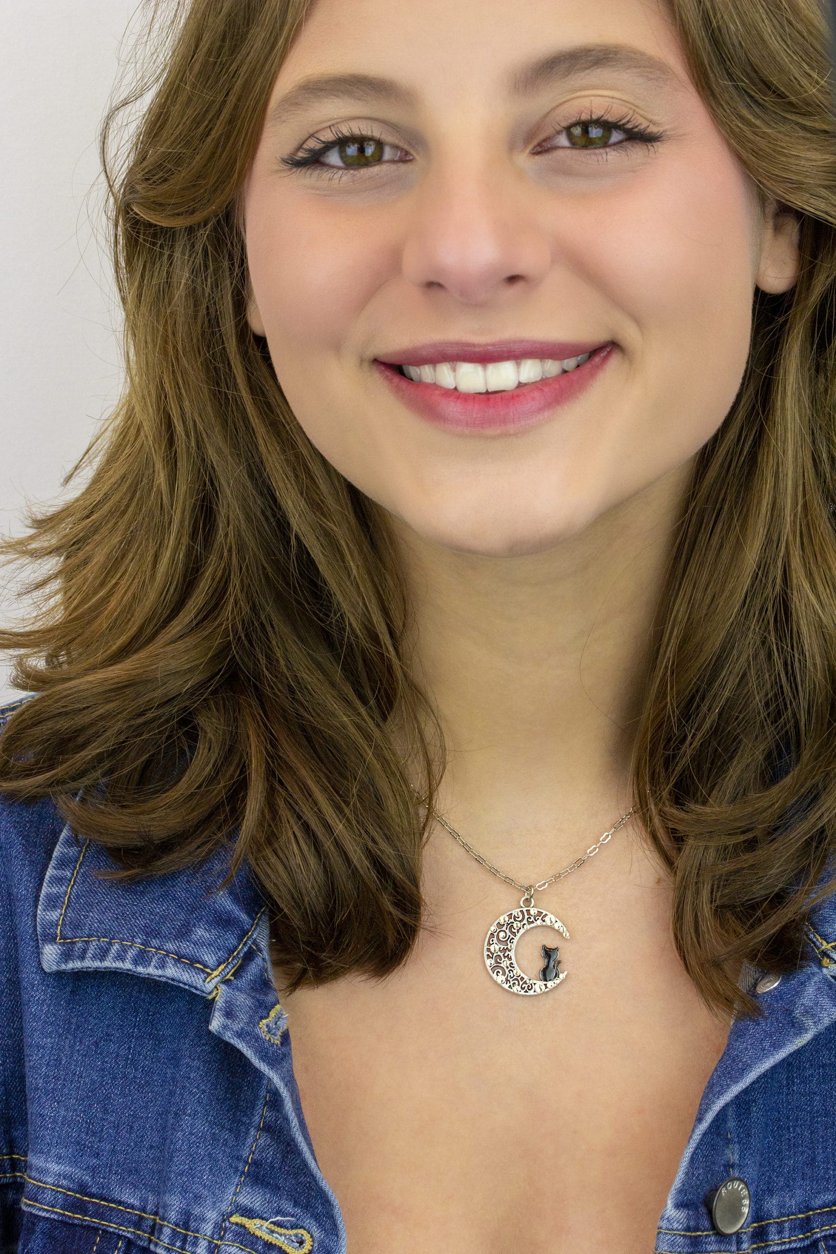 A smiling woman with wavy brown hair wears a denim jacket and the Crescent Moon w/Black Cat Necklace by Anne Koplik Designs, Inc., standing against a plain light background.