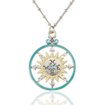 The NK: Round Turquoise Ring Yellow Sun Compass by Anne Koplik Designs, Inc. features a round compass pendant with a yellow sun design, light blue accents, and directional letters on a beaded chain, shown reflected on a white surface.