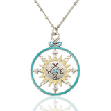 The NK: Round Turquoise Ring Yellow Sun Compass by Anne Koplik Designs, Inc. features a round compass pendant with a yellow sun design, light blue accents, and directional letters on a beaded chain, shown reflected on a white surface.