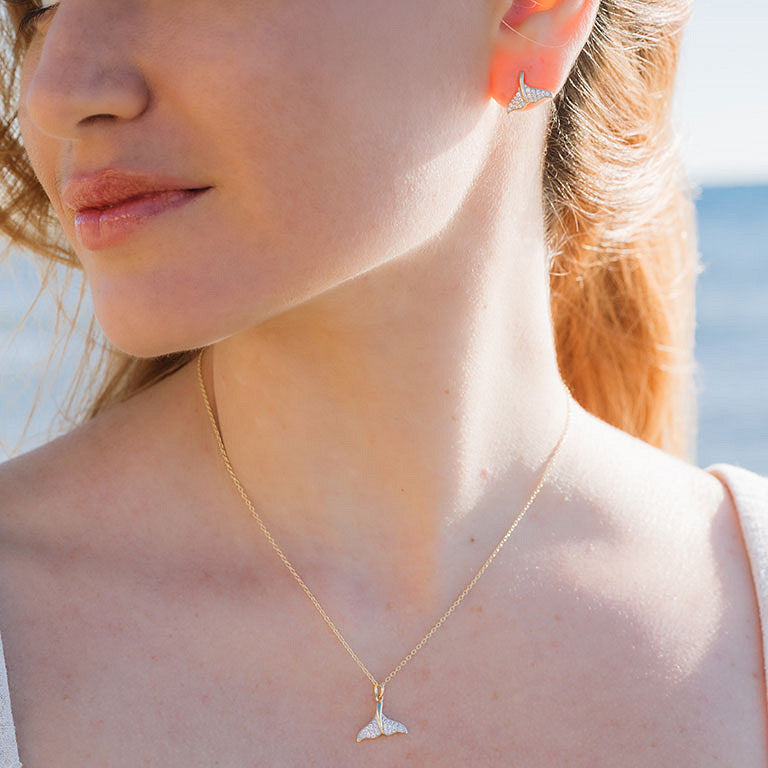 A woman wears the Shanore Gold Vermeil & White Sapphire Whale Tail Mini Necklace with matching stud earrings, standing outdoors against a blurred water and sky backdrop. The necklace sparkles with delicate white sapphires.