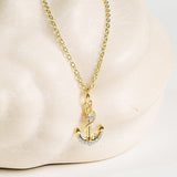 The Shanore Gold Vermeil & White Sapphire Anchor Mini Necklace features a dainty anchor pendant adorned with white sapphires, showcased on a pale, textured background.