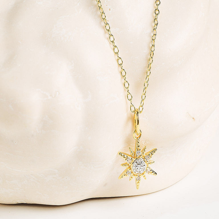 Shanore’s Gold Vermeil & White Sapphire Sun Mini Necklace is shown with its sun-shaped pendant against a cream, textured backdrop.