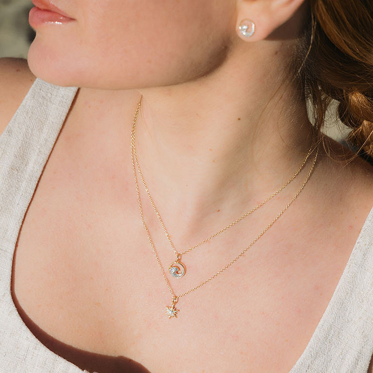 A woman in a cream sleeveless top wears the Shanore Gold Vermeil & White Sapphire Sun Mini Necklace layered with another necklace and small round stud earrings for a delicate, sparkling look.
