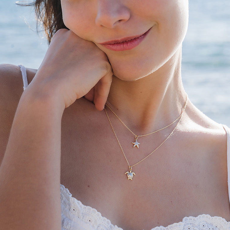 A woman in a white lace top smiles gently, her chin resting on her hand. She layers gold necklaces, including Shanore’s Gold Vermeil & White Sapphire Turtle Mini Necklace. The blurred ocean serves as a serene backdrop.
