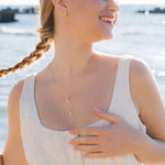 A woman with a braided hairstyle smiles by the water, sunlight catching her Shanore Gold Vermeil & White Sapphire Wave Mini Necklace as she enjoys the breeze in a sleeveless linen dress and layered gold jewelry against a softly blurred backdrop.