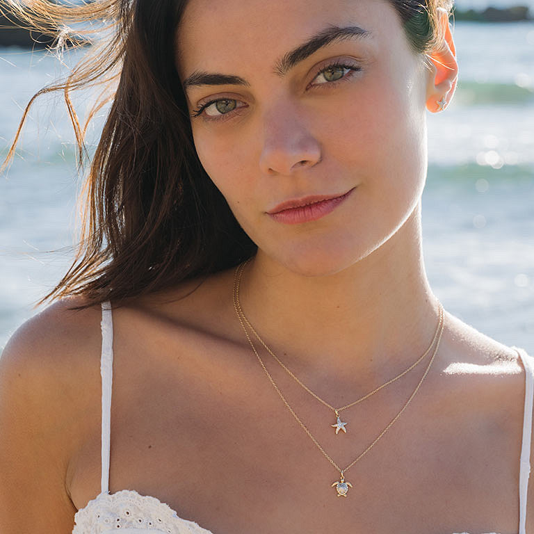 A woman in a white spaghetti-strap top wears the Shanore Gold Vermeil & White Sapphire Starfish Mini Necklace as she stands outdoors, sunlight brightening her face with the ocean sparkling in the background.
