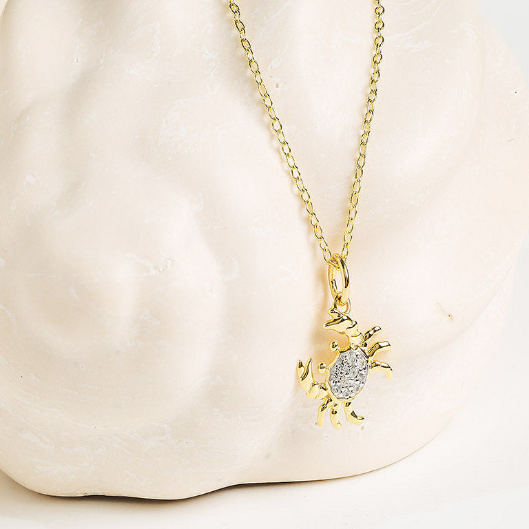 The Shanore Gold Vermeil & White Sapphire Crab Mini Necklace, featuring a crab-shaped pendant with white sapphires, is shown against a smooth, light-colored stone background.