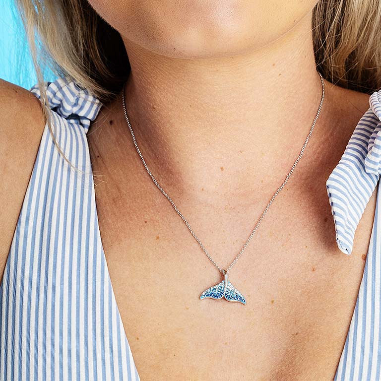 A woman wears a blue-and-white striped sleeveless top and a Shanore Swarovski Crystals Blue/White Crystal Whale Tail Necklace. The background is light blue.