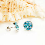 A close-up showcases Shanores Swarovski Crystals Aquamarine Round Stud Earrings from their Ocean Jewelry collection, placed in front of a textured seashell on a white surface. The earrings sparkle under soft light, with blue and white stones evoking aquamarine elegance.