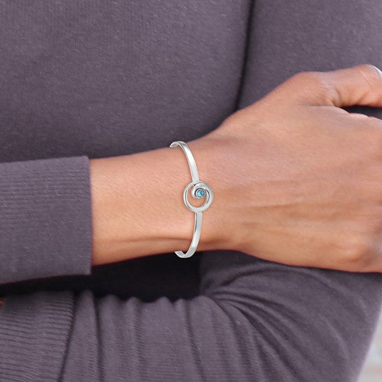 A person wearing the Presco/Quality Gold Crystal Wave Bangle Bracelet, a simple sterling silver piece featuring a circular design with a small blue gemstone at the center, shown against a long-sleeve dark top.