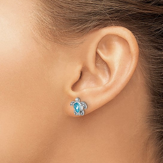 A womans ear is shown wearing Presco/Quality Golds Swarovski Crystals Turtle/Aquamarine Stud Earrings, crafted in sterling silver with a sparkling blue center. Her pulled-back hair highlights the unique turtle-shaped crystal studs.