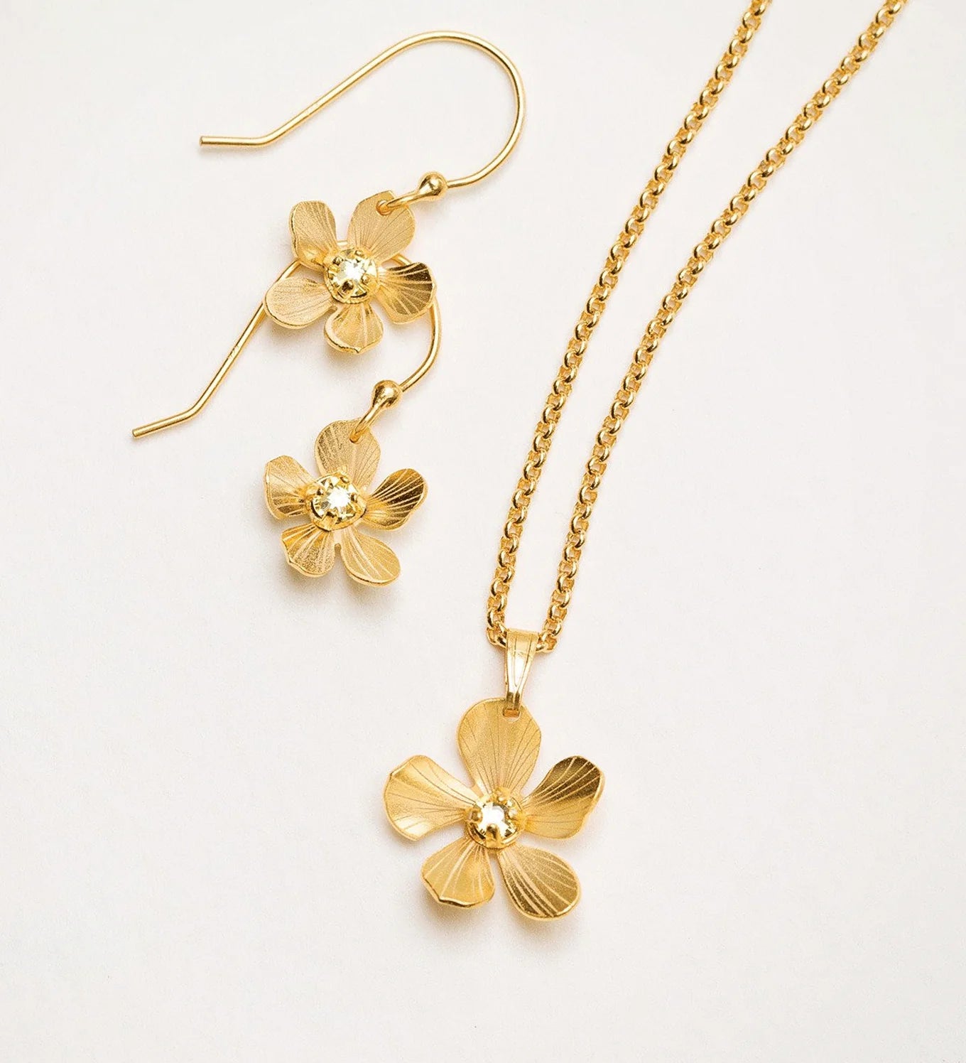 The Plumeria Drop Gold Necklace by Holly Yashi features hand-formed five-petal blooms with a textured finish, delicate chain, and gold bead centers, displayed with matching earrings from Holly Yashi Jewelry.