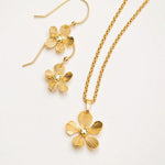 The Plumeria Drop Gold Necklace by Holly Yashi features hand-formed five-petal blooms with a textured finish, delicate chain, and gold bead centers, displayed with matching earrings from Holly Yashi Jewelry.