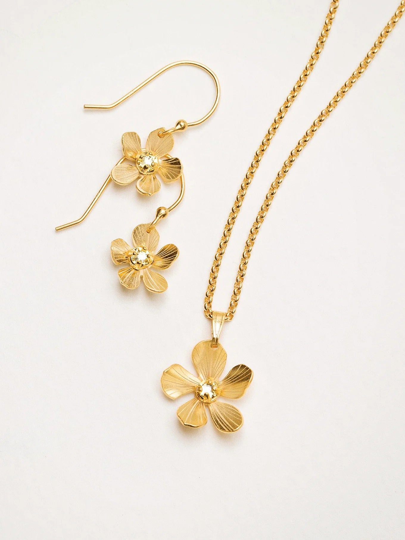 The Plumeria Drop Gold Necklace by Holly Yashi features hand-formed five-petal blooms with a textured finish, delicate chain, and gold bead centers, displayed with matching earrings from Holly Yashi Jewelry.