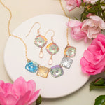 The Flora Multi Color Necklace by Holly Yashi, featuring vibrant floral-patterned square pendants, is displayed on a white round plate with pink flowers and green leaves—a stunning handcrafted jewelry set.