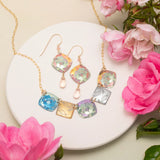 The Flora Multi Color Necklace by Holly Yashi, featuring vibrant floral-patterned square pendants, is displayed on a white round plate with pink flowers and green leaves—a stunning handcrafted jewelry set.