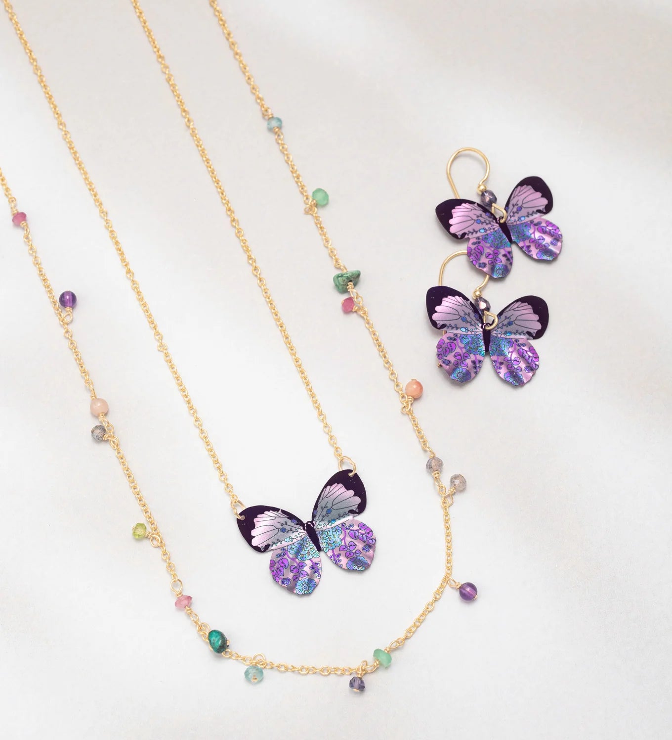 The Holly Yashi Blooming Butterfly Radiant Purple Necklace features an iridescent niobium butterfly pendant with matching earrings and multicolored beads, shown on white. Some pieces offer adjustable gold chains for a perfect fit.