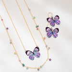 The Holly Yashi Blooming Butterfly Radiant Purple Necklace features an iridescent niobium butterfly pendant with matching earrings and multicolored beads, shown on white. Some pieces offer adjustable gold chains for a perfect fit.