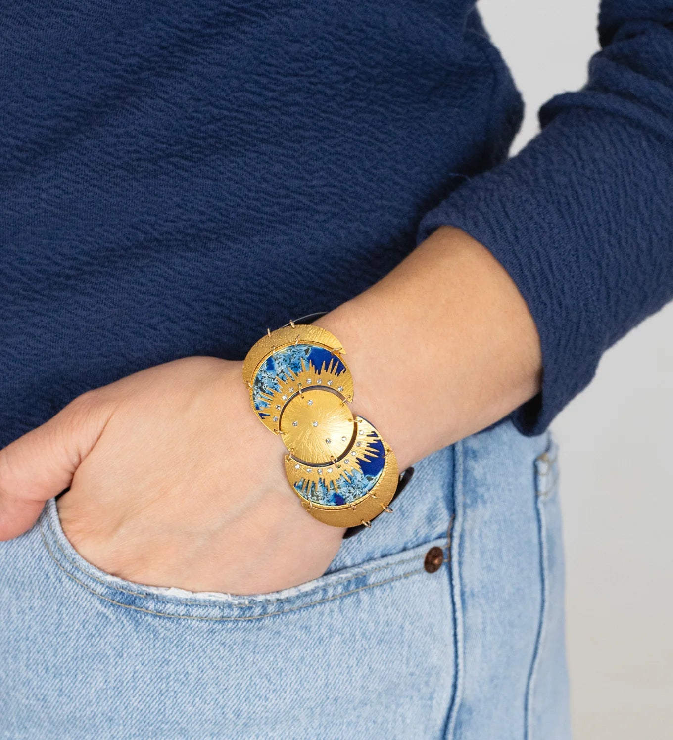 A person in light blue jeans and a navy sweater has their hand in their pocket, highlighting the Holly Yashi Selena Midnight Blue Bracelet—an elegant gold piece with sunburst motifs that brings celestial charm to their wrist.