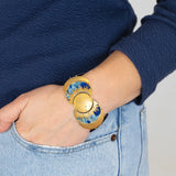 A person in light blue jeans and a navy sweater has their hand in their pocket, highlighting the Holly Yashi Selena Midnight Blue Bracelet—an elegant gold piece with sunburst motifs that brings celestial charm to their wrist.
