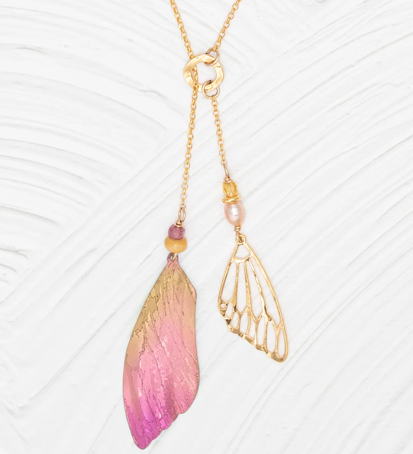 The Special Edition Flutterby Pink Lariat Necklace by Holly Yashi features an asymmetrical design: one side boasts a pink and gold ombré wing charm with beads, the other a delicate openwork gold wing with pearl, all set on an adjustable chain.