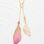The Special Edition Flutterby Pink Lariat Necklace by Holly Yashi features an asymmetrical design: one side boasts a pink and gold ombré wing charm with beads, the other a delicate openwork gold wing with pearl, all set on an adjustable chain.