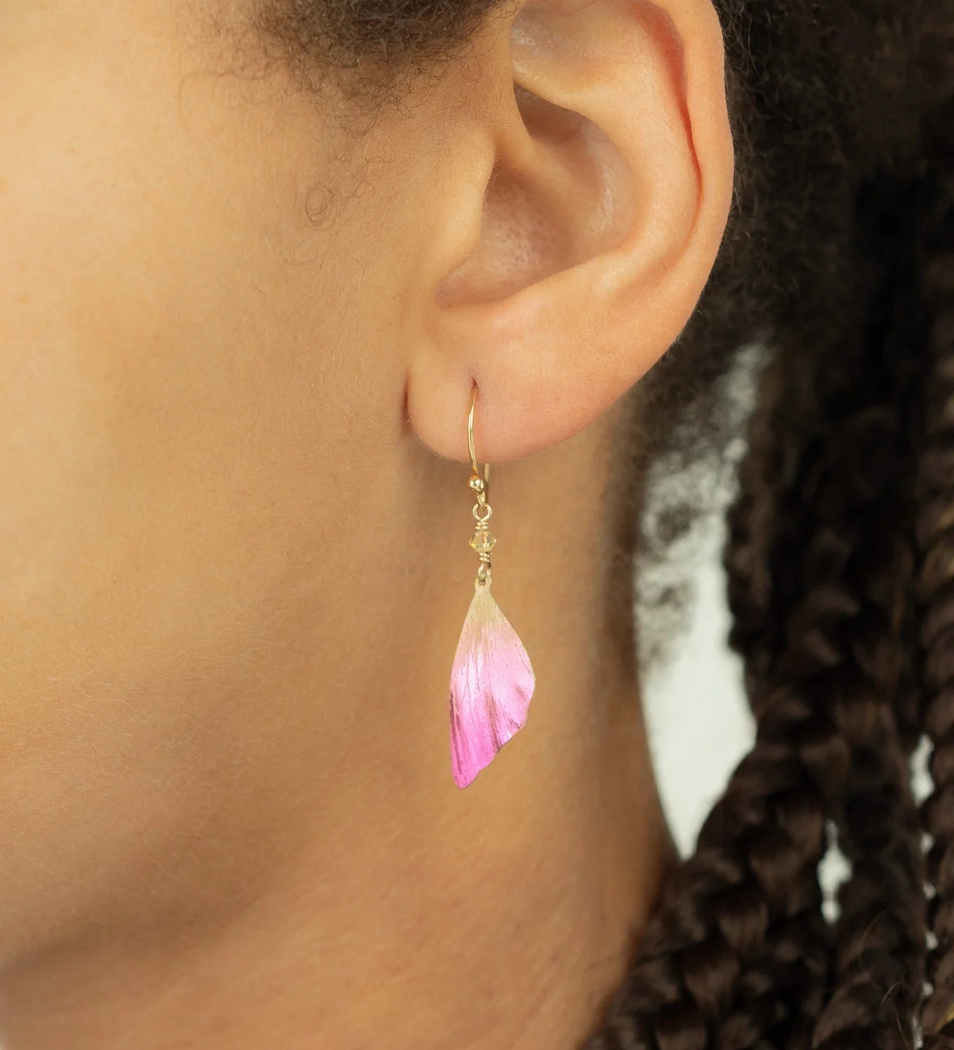 Close-up of a person’s ear wearing Special Edition Flutterby Pink Earrings by Holly Yashi, featuring a gold hook and pink and white petal-shaped charm. The earrings stand out beautifully against their braided hair, symbolizing cancer awareness.