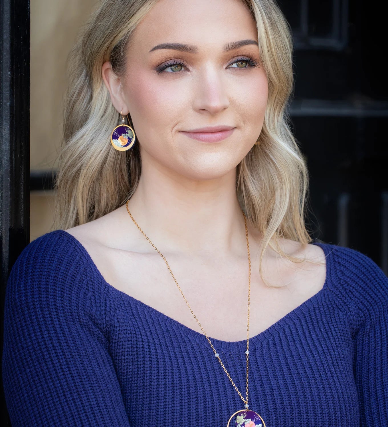 A woman with long blonde hair smiles softly in front of a dark background, wearing a blue ribbed top and Luna Earrings/Blue Moon by Holly Yashi—circular pendants featuring colorful abstract designs.