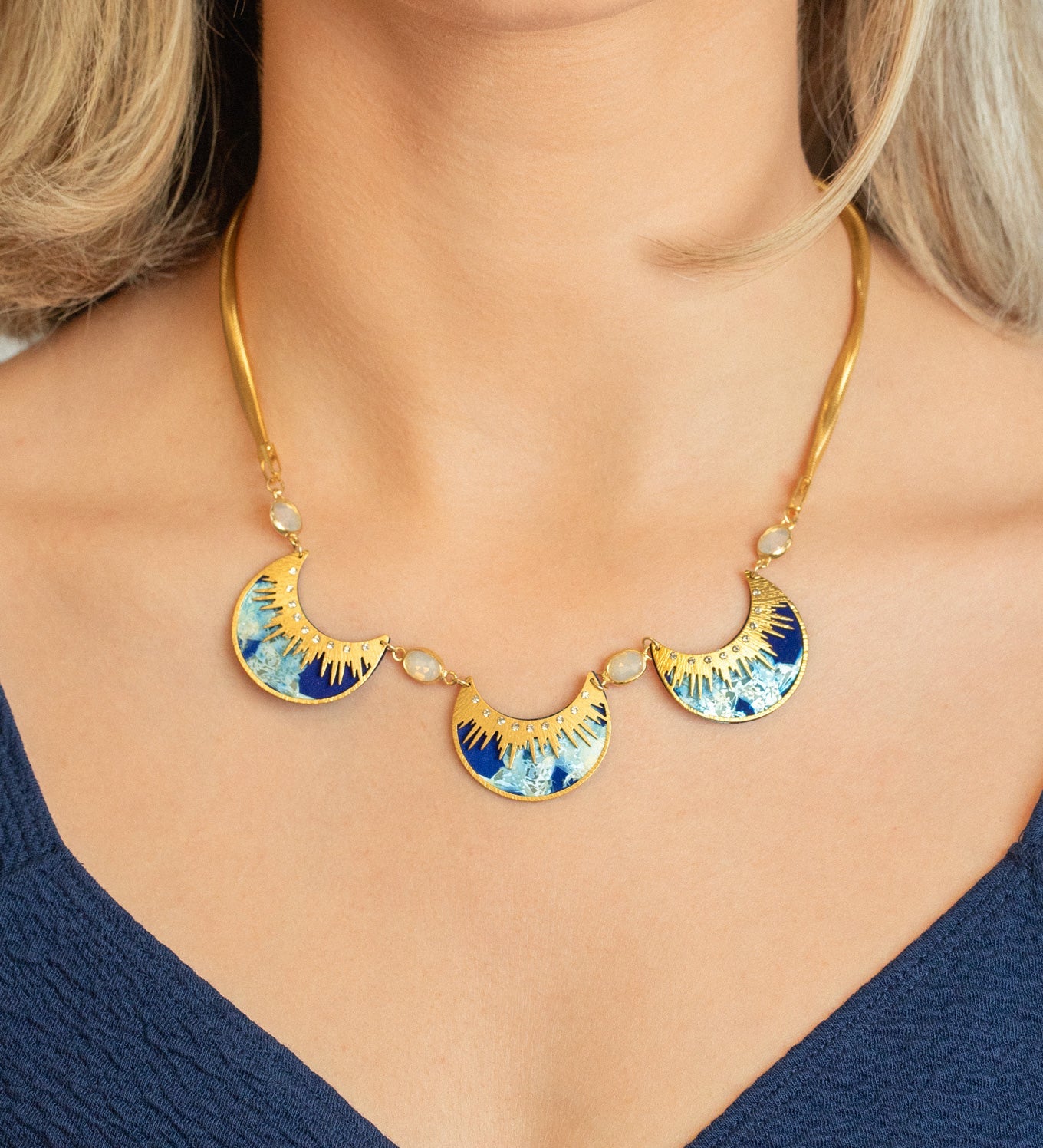 A woman wears the Holly Yashi Selena Necklace in Midnight Blue, featuring three sunburst-patterned blue and gold fan pendants, paired with a deep blue V-neck top.