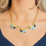 A woman wears the Holly Yashi Selena Necklace in Midnight Blue, featuring three sunburst-patterned blue and gold fan pendants, paired with a deep blue V-neck top.