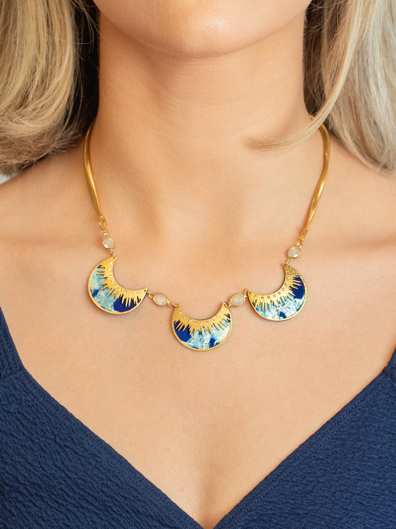 A woman wears the Holly Yashi Selena Necklace in Midnight Blue, featuring three sunburst-patterned blue and gold fan pendants, paired with a deep blue V-neck top.