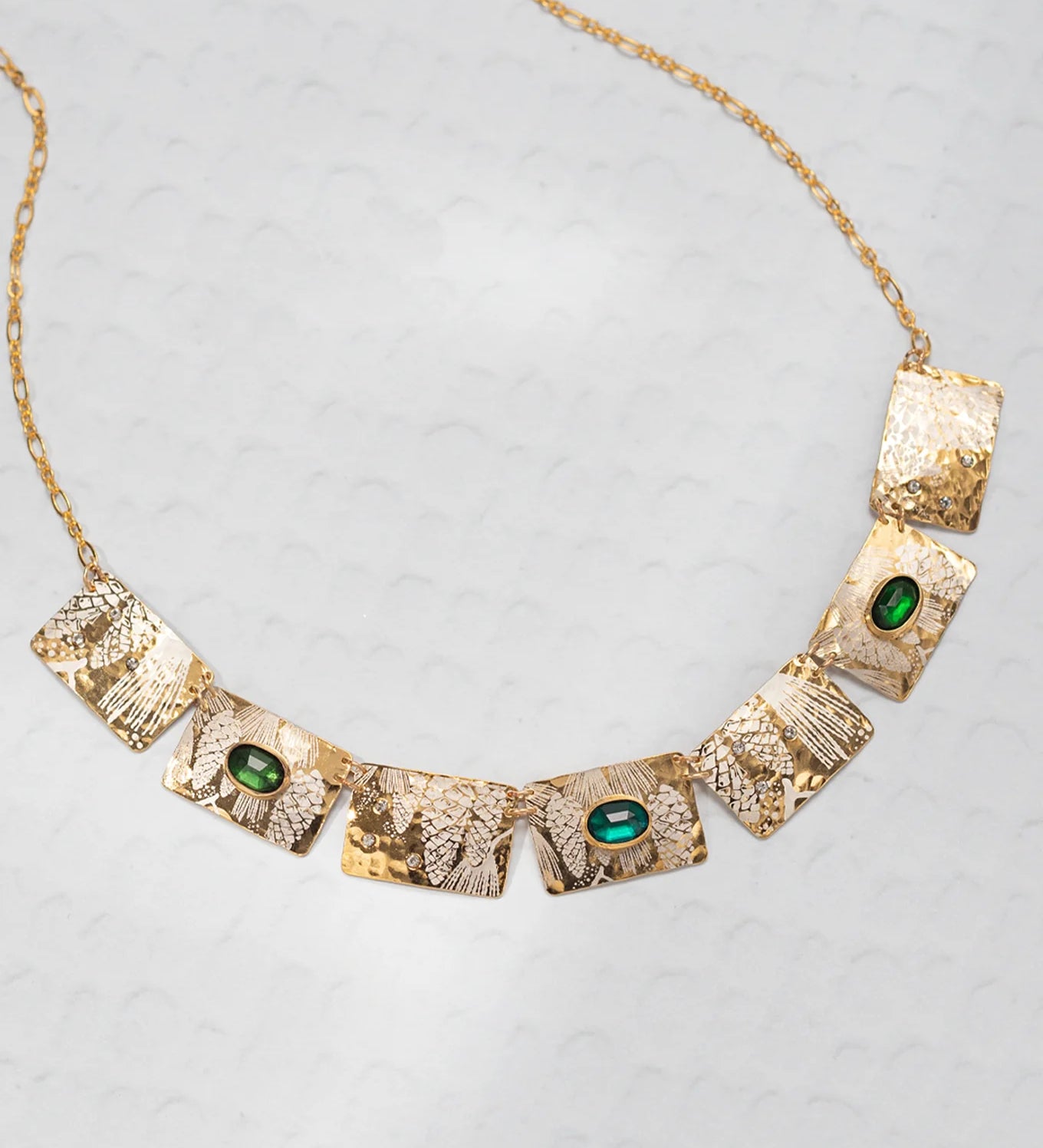 The Gilded Petra Emerald Necklace by Holly Yashi showcases seven hammered rectangles, three with emerald green crystals, on a delicate gold chain. This Victorian-inspired piece shines with elegance on a light textured backdrop.