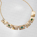 The Gilded Petra Emerald Necklace by Holly Yashi showcases seven hammered rectangles, three with emerald green crystals, on a delicate gold chain. This Victorian-inspired piece shines with elegance on a light textured backdrop.